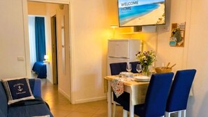 A hotel room with a dining area, a television displaying a beach scene, a ceiling fan, and a bed with a blue cover. - "CASA POMONTE" ON THE SEA with PRIVATE PARKING SPACE. (Pomonte)