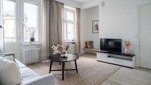 A modern living room with a sofa, a TV on a stand, a round table with decorative items, and large windows with curtains. - 1BR Penthouse with Sauna & Private Rooftop Terrace (Tampere)