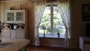 Una habitación con techo de madera, una ventana con cortinas florales, un armario blanco, una mesa de comedor con sillas y una pequeña planta en maceta en el alféizar.