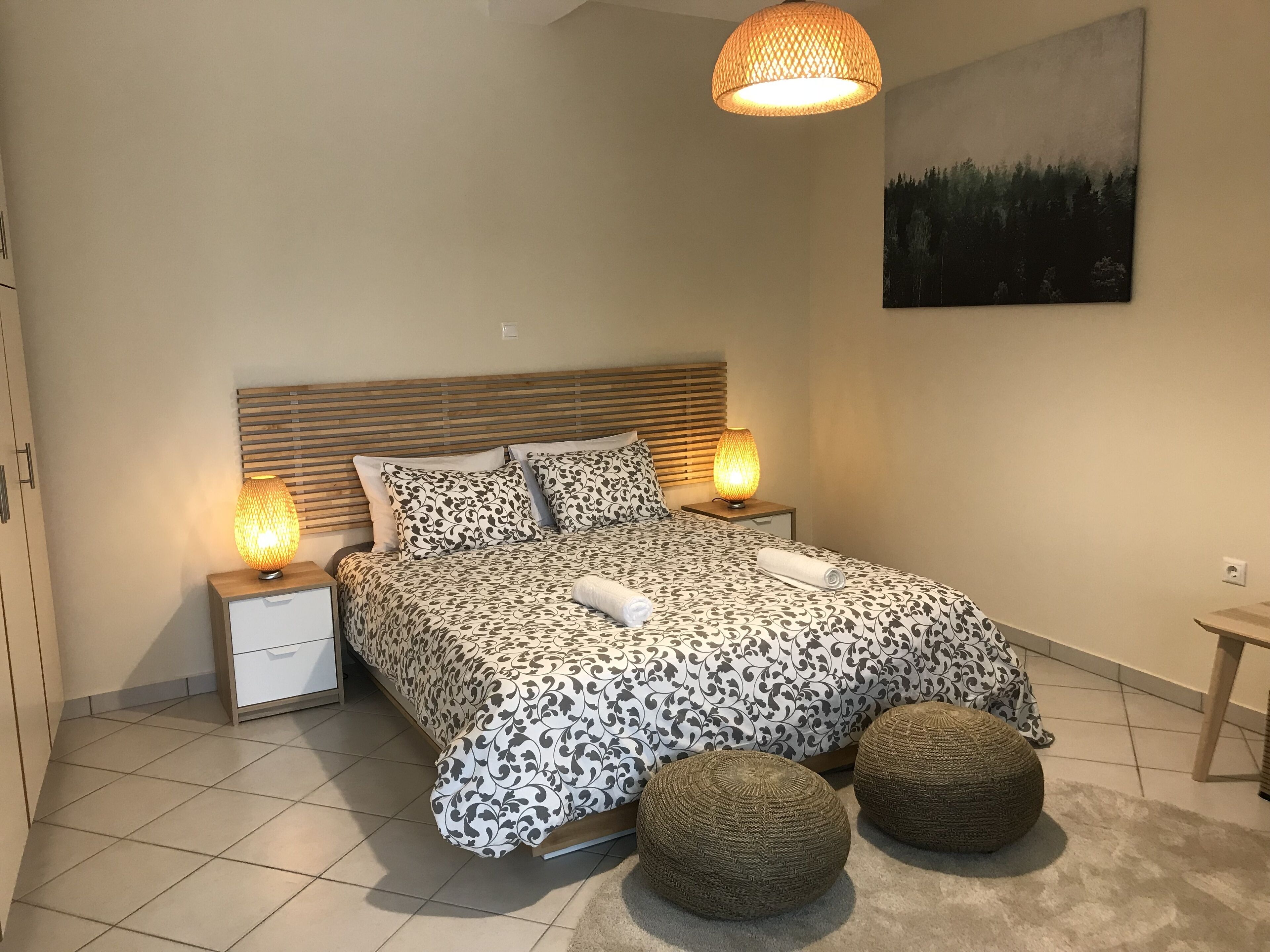 Ein ordentlich eingerichtetes Schlafzimmer mit einem Bett, zwei Nachttischlampen, einem Kopfteil, einem an der Wand hängenden Gemälde und zwei runden Hocker.