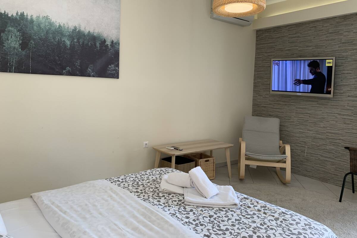 Habitación de hotel con una cama, un televisor montado en la pared, una silla, una mesita y un cuadro en la pared.