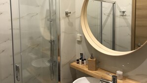 A modern bathroom with a glass-enclosed shower, a round mirror, and a wooden vanity with toiletries. - Flight Apartment Airport (Spata-Artemida)