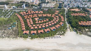 An aerial view of a coastal resort with a circular layout of villas, beachfront access, and a central pool area.