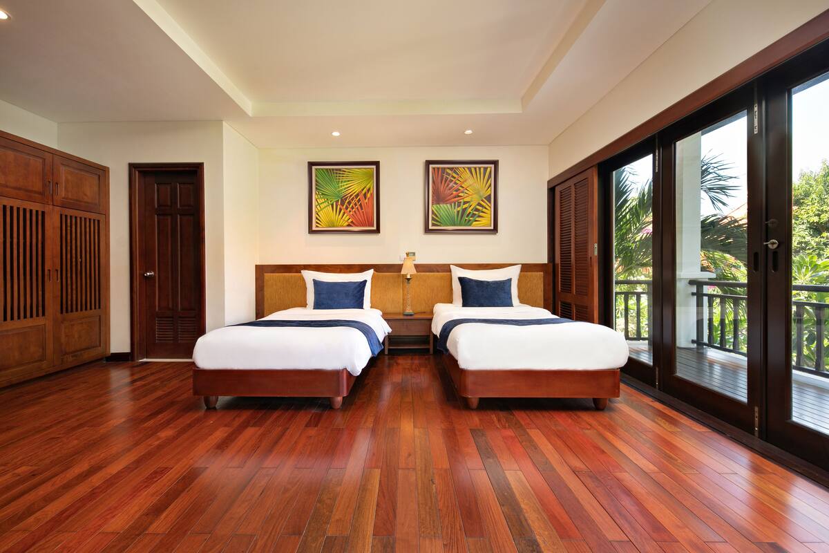 A hotel room with two beds, wooden flooring, a large window, and two framed pictures on the wall.