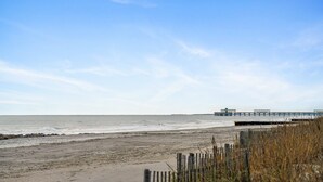 Une plage avec une jetée en bois qui s’avance dans l’eau, un rivage sablonneux et un ciel dégagé.