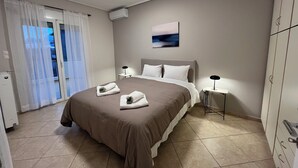 A bedroom with a bed, two bedside lamps, a window with curtains, an air conditioning unit, and a painting on the wall.
