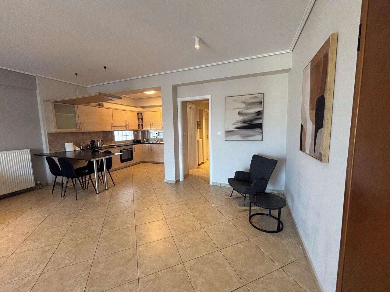 A modern kitchen with a dining area, a hallway, and a living space with a chair and artwork.
