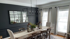 Un comedor con mesa de madera, sillas blancas, una lámpara de araña y un espejo grande en la pared.