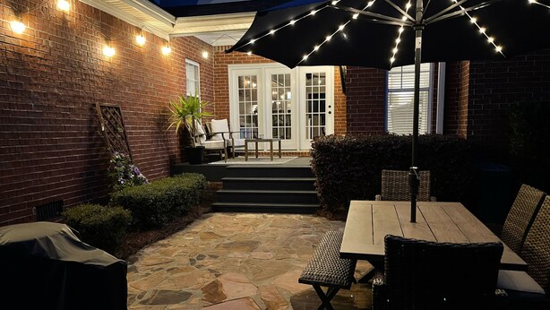 Un patio exterior con suelo de piedra, una mesa y sillas, y luces colgantes.