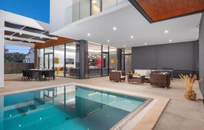 Un espace piscine extérieur moderne doté d’un espace repas clos par des baies vitrées, de coins salon extérieurs et offrant une vue sur le bâtiment adjacent.