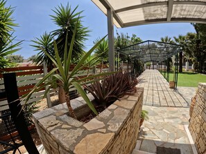 A covered walkway with stone flooring, surrounded by various plants and a metal fence. - Hector Villa Airport (Spata-Artemida)