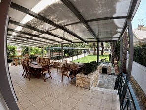 A covered patio with a dining table and chairs, a stone planter, and a view of a green lawn and trees.