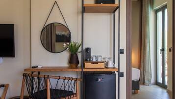 Een kleine kamer met een houten bureau, een kleine zwarte koelkast, een rieten stoel, een spiegel en een potplant.