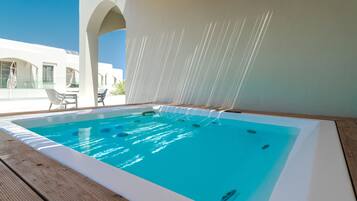 Una piscina con muelle de madera, pared blanca con fuente de agua, y un edificio con arcos al fondo.