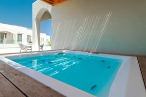 Una piscina con muelle de madera, pared blanca con fuente de agua, y un edificio con arcos al fondo.