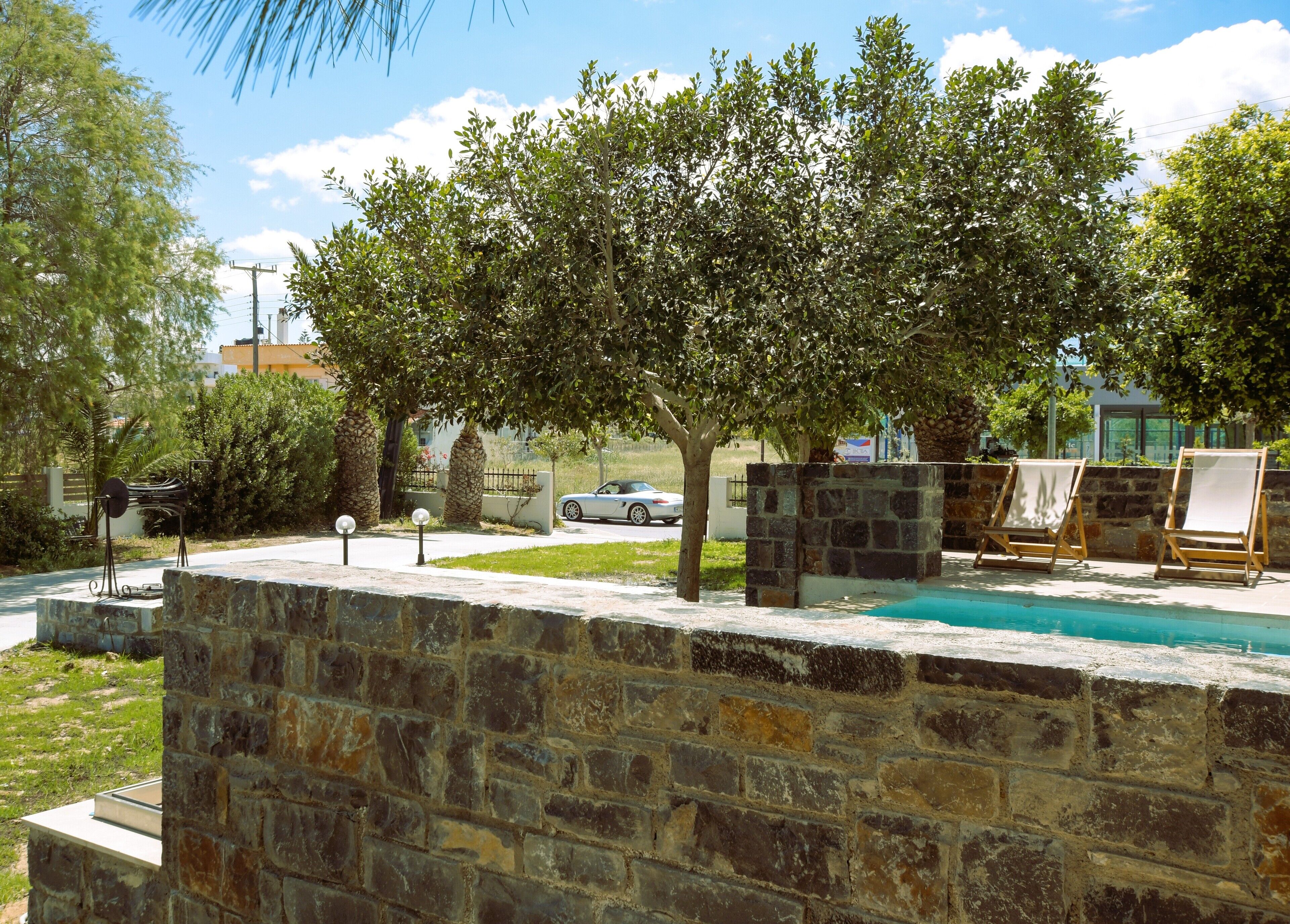 A stone wall with a pool, two wooden beach chairs, and a tree in a landscaped area.