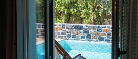 A glass door leading to a swimming pool with a stone wall and a wooden chair.