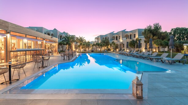 Piscina com espreguiçadeiras e mesas, rodeada por edifícios com janelas em arco e áreas de estar ao ar livre.