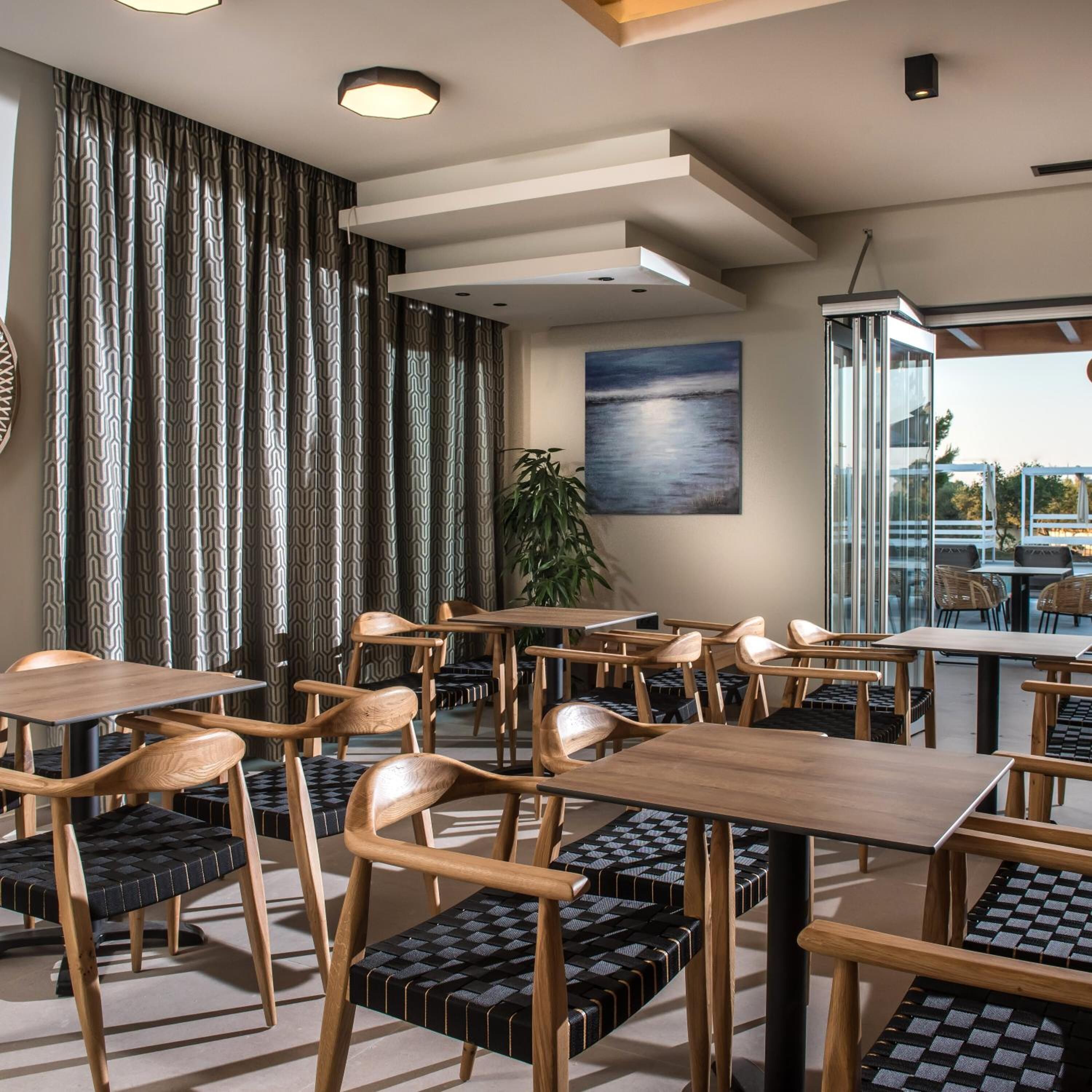 A modern restaurant with wooden tables and chairs, a large window with a view, and a painting on the wall.
