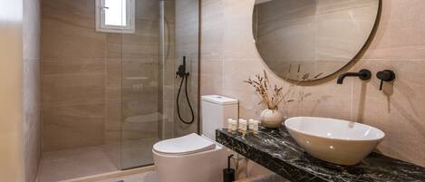 A modern bathroom with a glass-enclosed shower, a white toilet, a marble countertop, a round mirror, and a wooden shelf with towels.