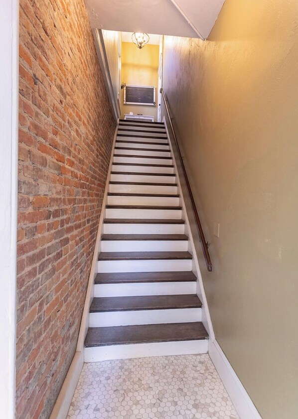 Una escalera con un muro de ladrillo a un lado y un muro liso al otro, que conduce a un rellano con una lámpara.
