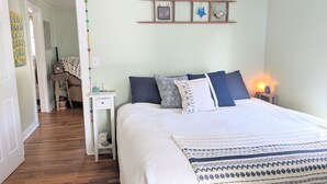 A bedroom with a bed, bedside tables, a nightstand, a wall-mounted shelf with decorative items, and a door leading to another room. - Cozy cottage with backyard firepit (Asheville)