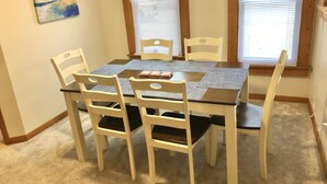 A dining area with a wooden table and six white chairs, a painting on the wall, and two windows with blinds. - South Wind, walk to the beach, zoo, & more! (Michigan City)
