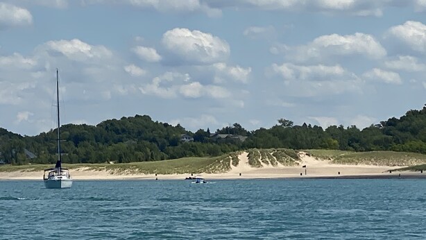 A sailboat on a calm sea with a beach and forested hills in the background. - South Wind, walk to the beach, zoo, & more! (Michigan City)