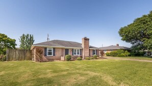 A single-story house with a brick chimney, a front yard with a wooden fence, and a clear sky. - Walk to Stadium! Modern Home + Free Parking (Tuscaloosa)