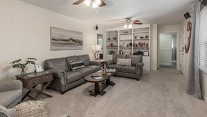 A living room with a grey sofa, a coffee table, and a bookshelf. There is a ceiling fan, a painting on the wall, and a window with curtains. - Walk to Stadium! Modern Home + Free Parking (Tuscaloosa)