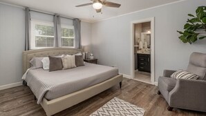 A bedroom with a bed, a ceiling fan, a chair, and a bathroom visible through an open door. - Walk to Stadium! Modern Home + Free Parking (Tuscaloosa)