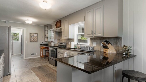 A modern kitchen with grey cabinets, a central island, stainless steel appliances, and a bar stool. - Walk to Stadium! Modern Home + Free Parking (Tuscaloosa)