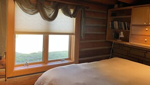 Quarto de cabana de madeira com cama, janela com cortinas, estante e abajur.