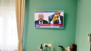A room with a TV displaying a news broadcast, a wooden desk, a white chair, and a window with curtains. - Villa Bellavista (Alba)
