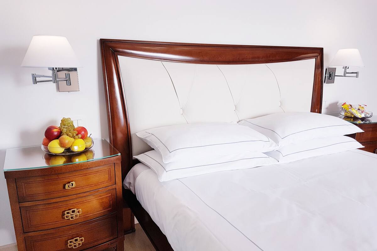 A neatly made bed with white linens, a wooden nightstand with drawers, and a fruit bowl on the nightstand.