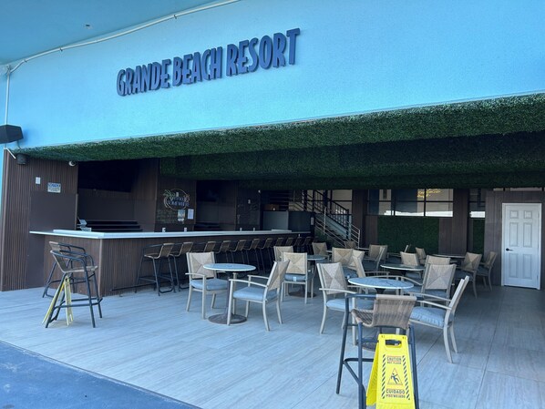 A Grande Beach Resort outdoor seating area with tables and chairs, a bar, and a staircase leading to an upper level.