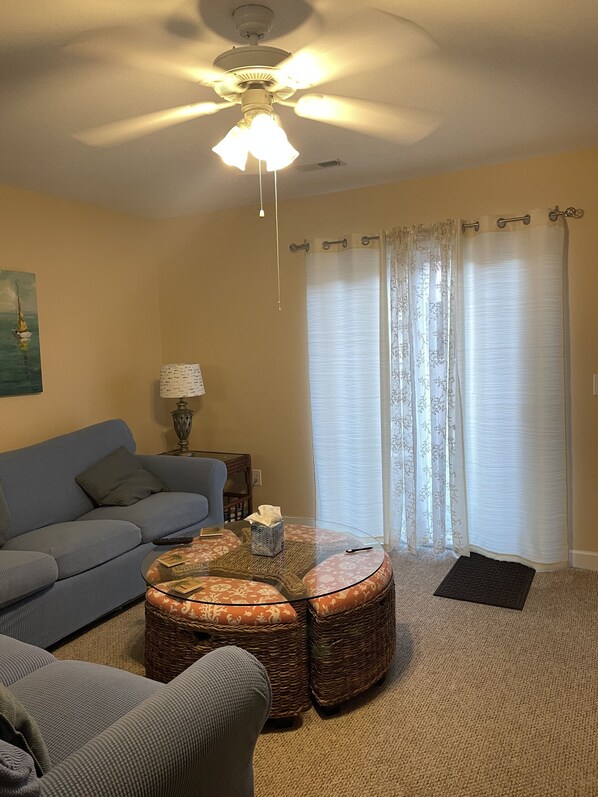 A living room with a blue sofa, a round coffee table with books, a ceiling fan, and a window with sheer curtains. - Absolutely Charming Firebird Townhome, 1 minute walk to Myrtle Beach! (Myrtle Beach)
