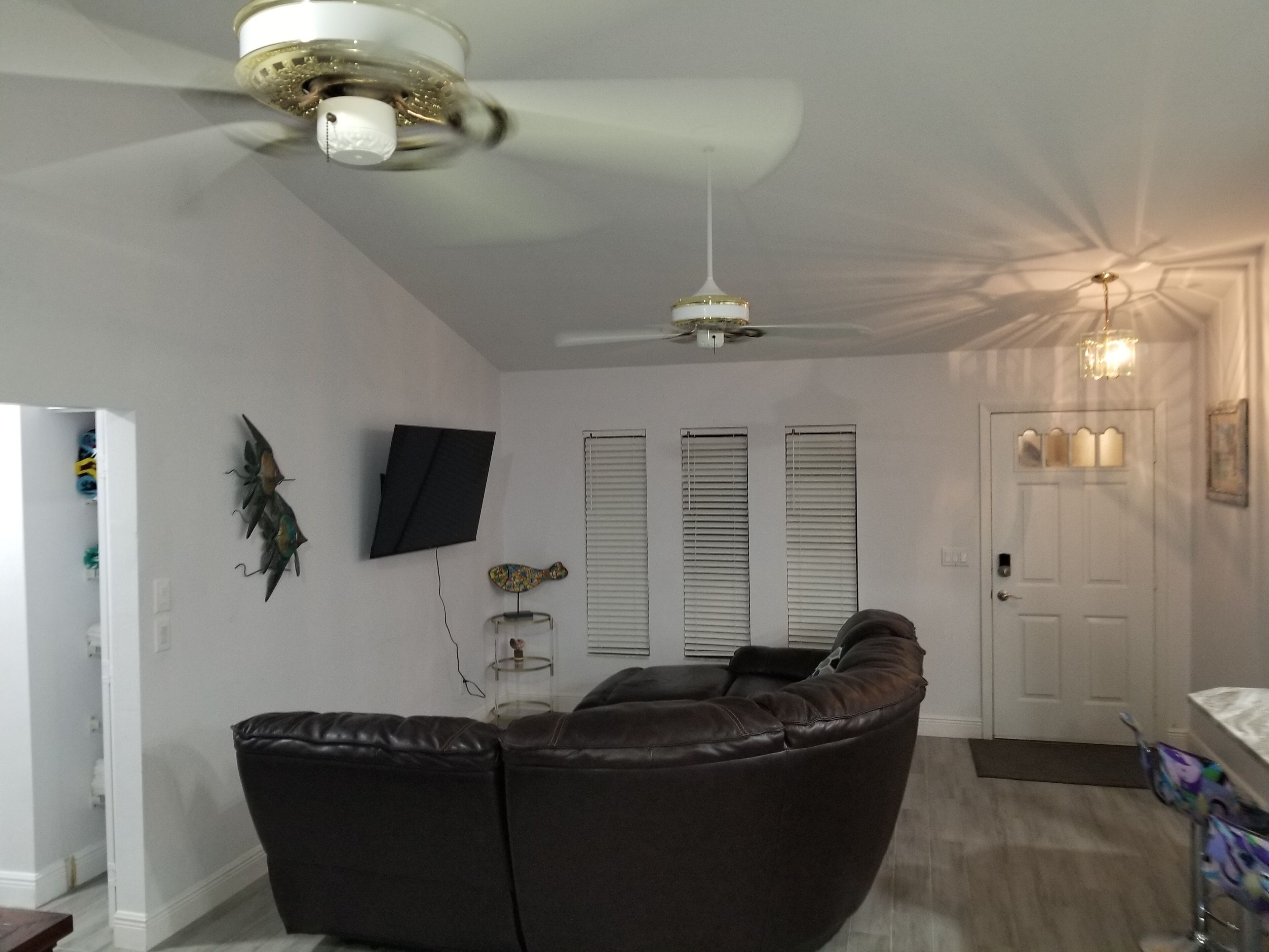 Un salon doté d’un canapé en cuir aux lignes courbes, d’un ventilateur de plafond, d’un téléviseur fixé au mur et d’une porte agrémentée d’un luminaire décoratif.