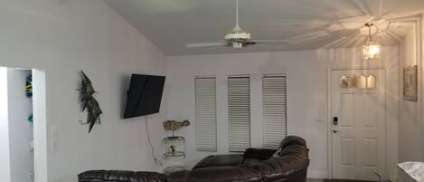 Un salon doté d’un canapé en cuir aux lignes courbes, d’un ventilateur de plafond, d’un téléviseur fixé au mur et d’une porte agrémentée d’un luminaire décoratif.
