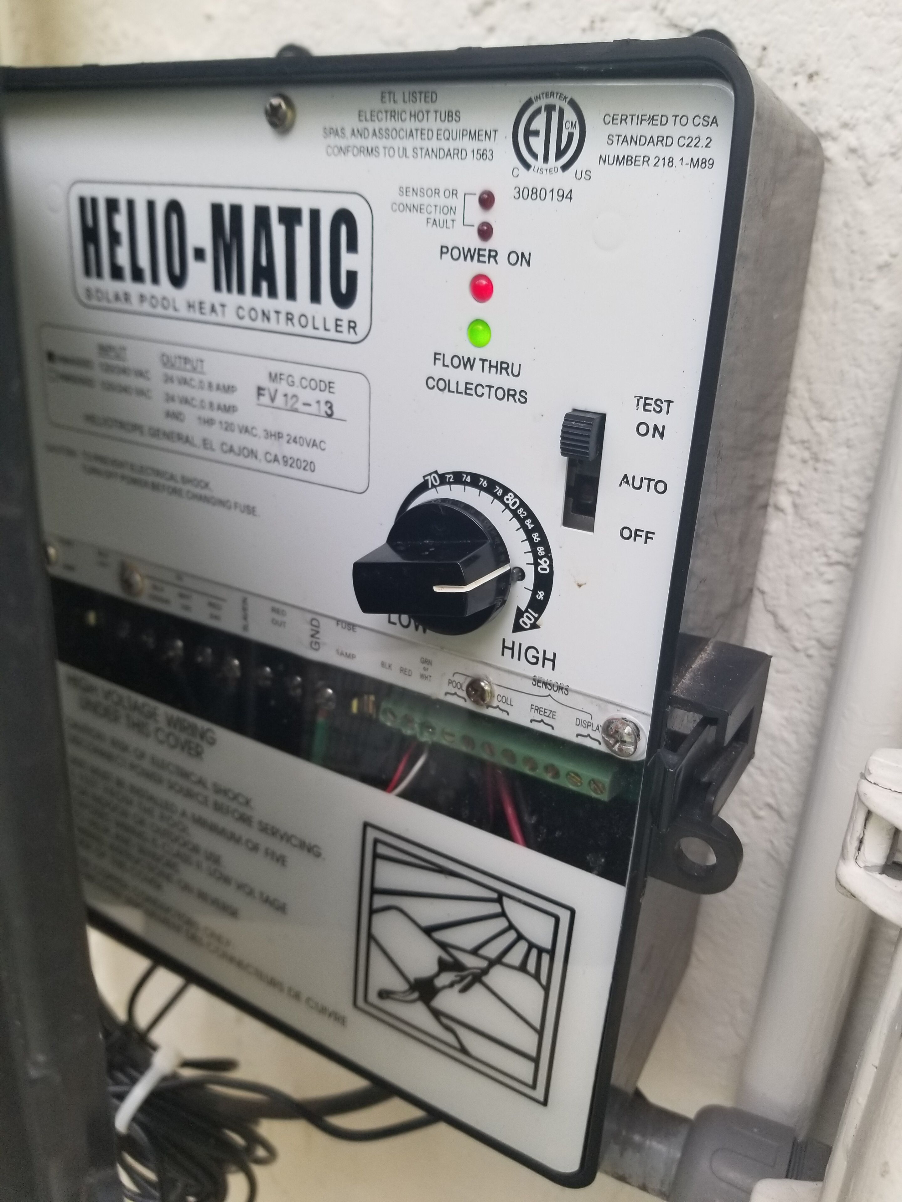 Controlador de calor per piscina amb forma helicoidal, un comandament giratori, botons de prova i un etiquetatge d'advertiment.