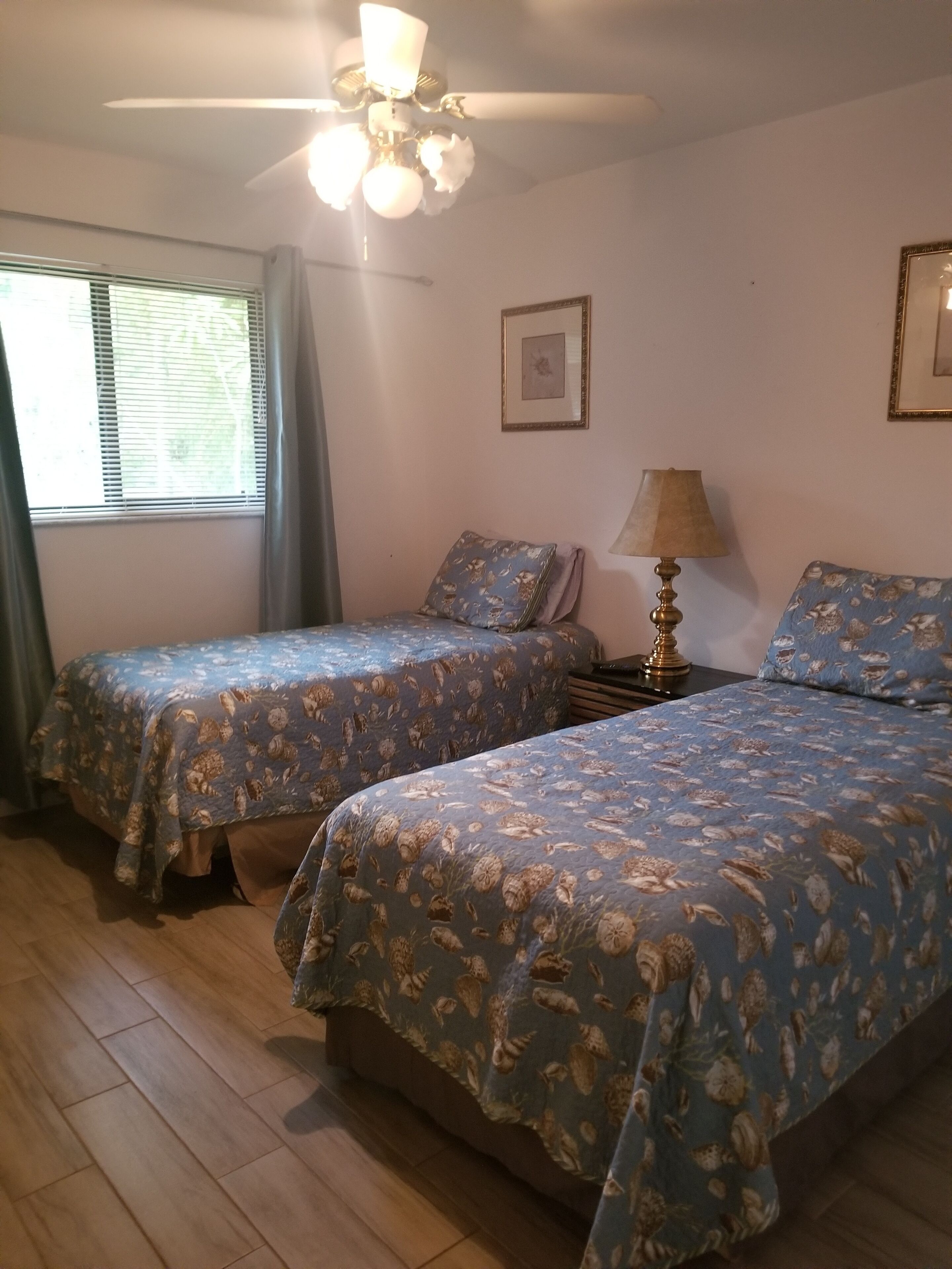 Un dormitorio con dos camas, un ventilador de techo, una ventana con persianas y un cuadro en la pared.