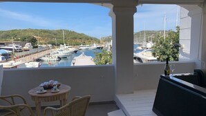 Een balkon met rieten stoelen en een tafeltje, met uitzicht op een jachthaven met boten en een glooiend landschap.