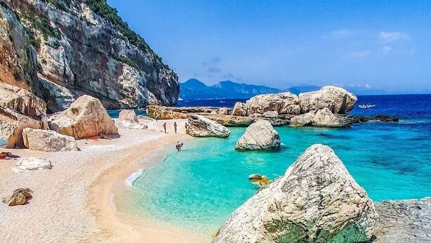 Een strand met helder turquoise water, grote rotsen en een rotsachtige klif.