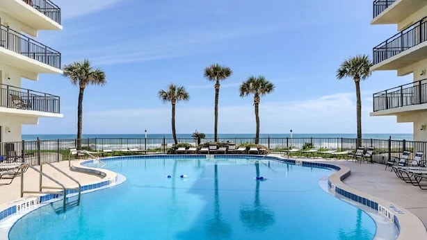 A swimming pool with a curved edge, surrounded by lounge chairs and palm trees, with a view of the ocean. - Ocean Front Condo in Ormond By The Sea ( Ormond Beach )  (Ormond Beach)