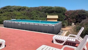 An outdoor swimming pool with a wooden structure in the background, surrounded by greenery and white plastic chairs.