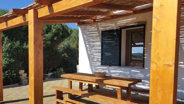 A wooden pergola structure with a picnic table and benches, a white building with a window, and a view of greenery and mountains in the background.