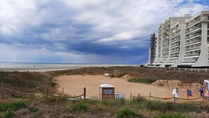 A beach view with a sandy area, a portable toilet, and a white building with multiple balconies. - l'Art de l'eau beach studio on private property (NEUFCHÂTEL-HARDELOT)