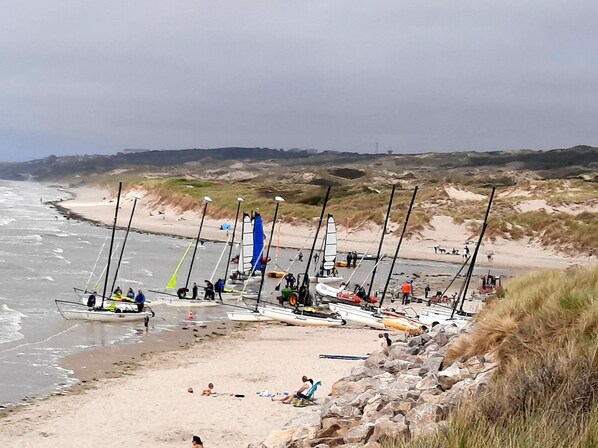 A beach with several boats docked near the shore, people walking, and a rocky landscape in the background.