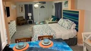 A cozy bedroom with a bed, bedside table, and a chair. There is a small table with a bowl and plates. The room has a window with curtains and a door.