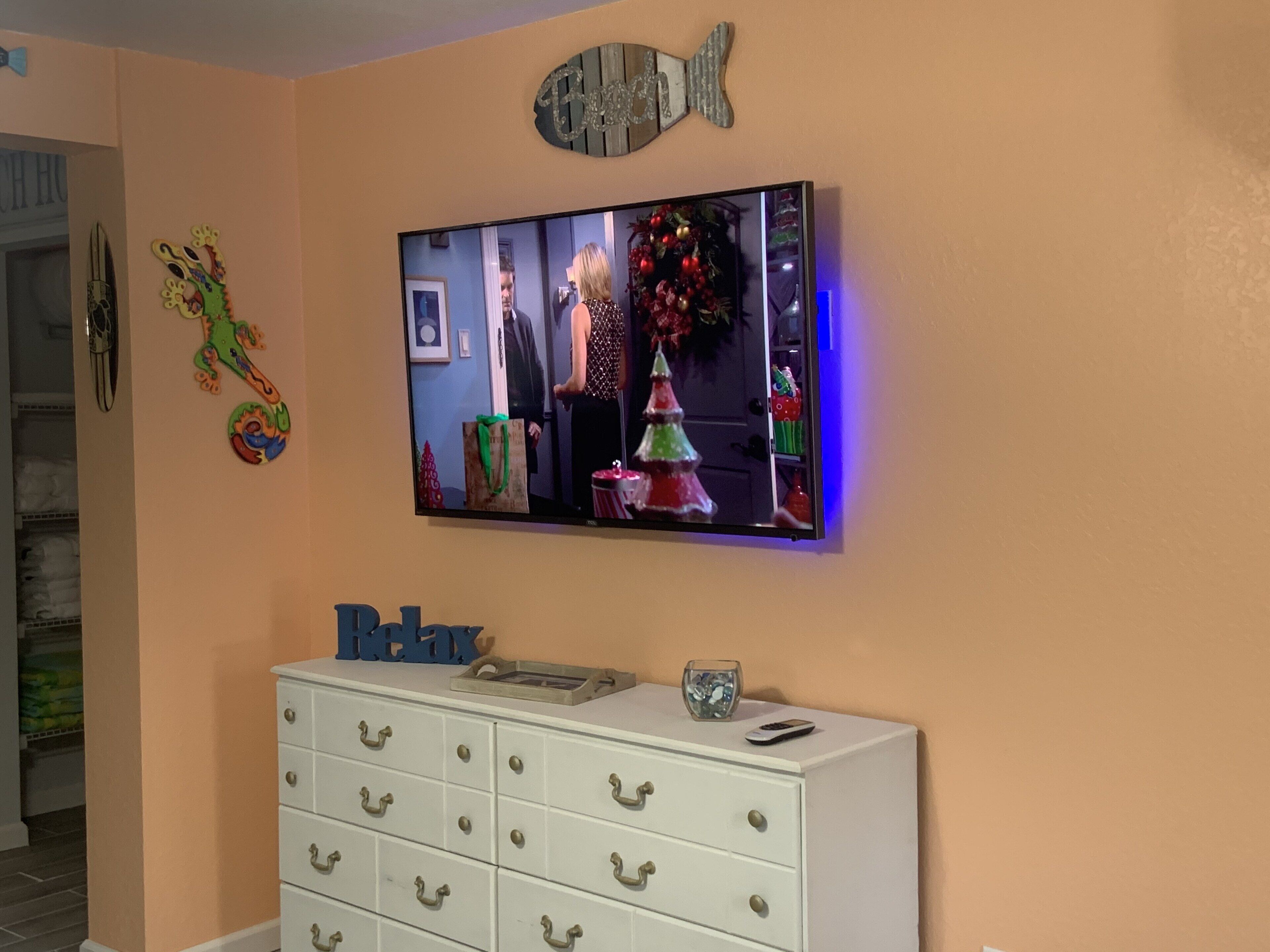 A living room with a television mounted on the wall, a white dresser with drawers, and a decorative fish-shaped mirror above the TV.